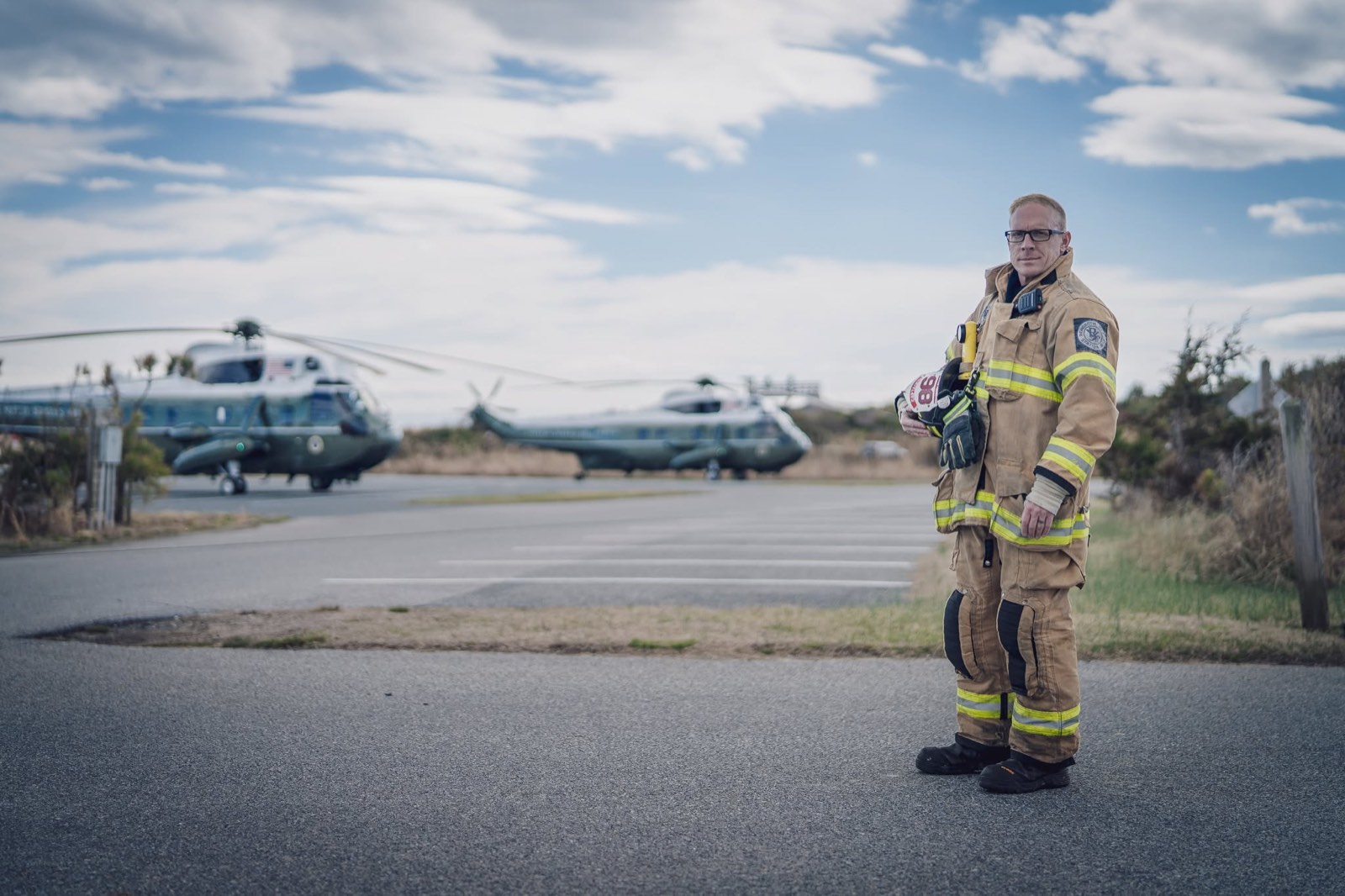 Ryan Peters in firefighter turnout gear holding his helmet, with Marine One on the pad behind him